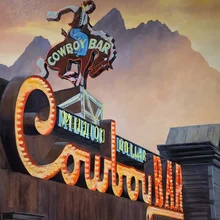 neon sign with a cowboy riding a bucking bronco and the words Cowboy Bar against a mountain backdrop at sunset